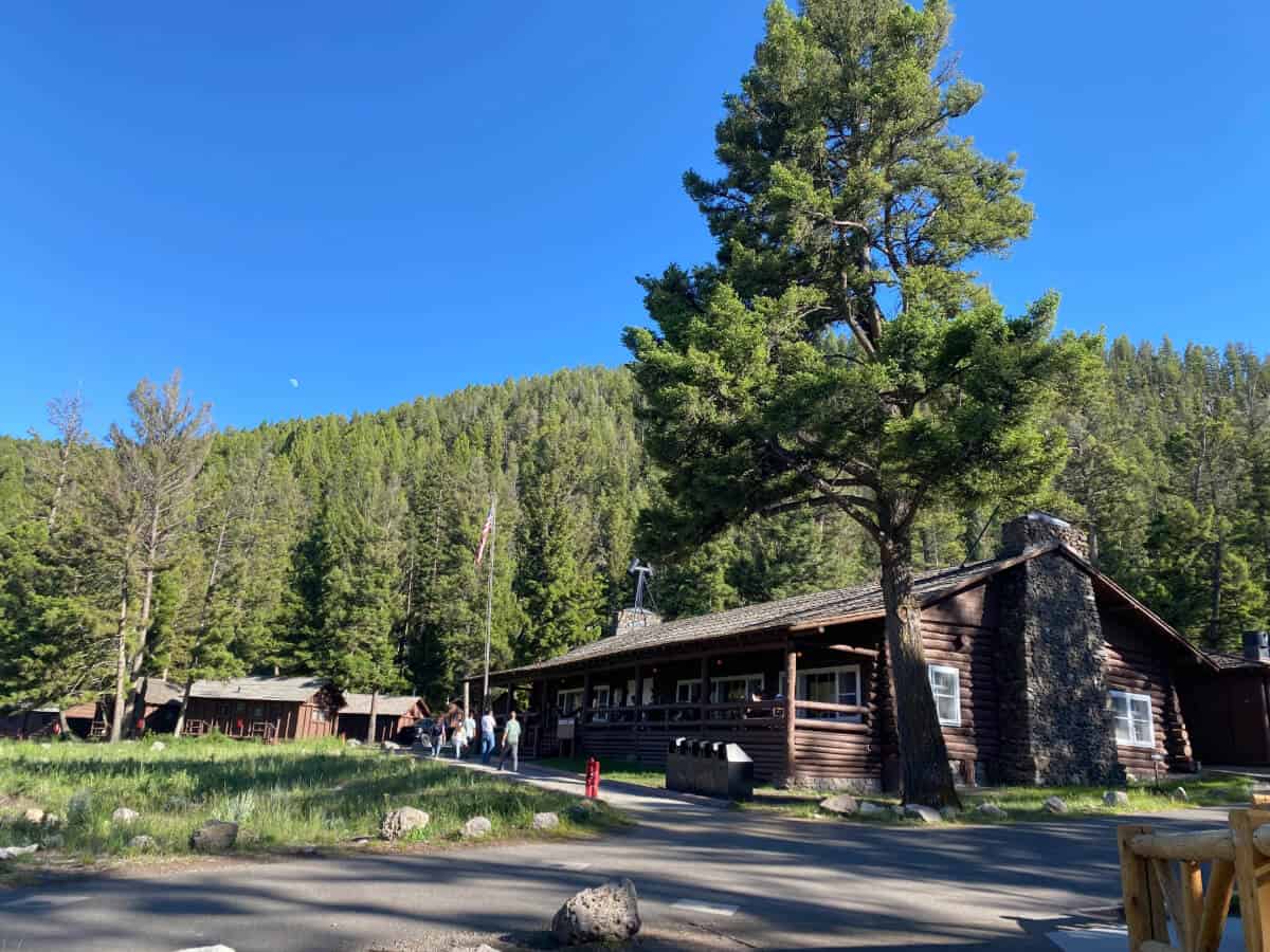 A long log cabin building with a stone chimney sits at the edge of a forested mountain slope under a clear blue sky.