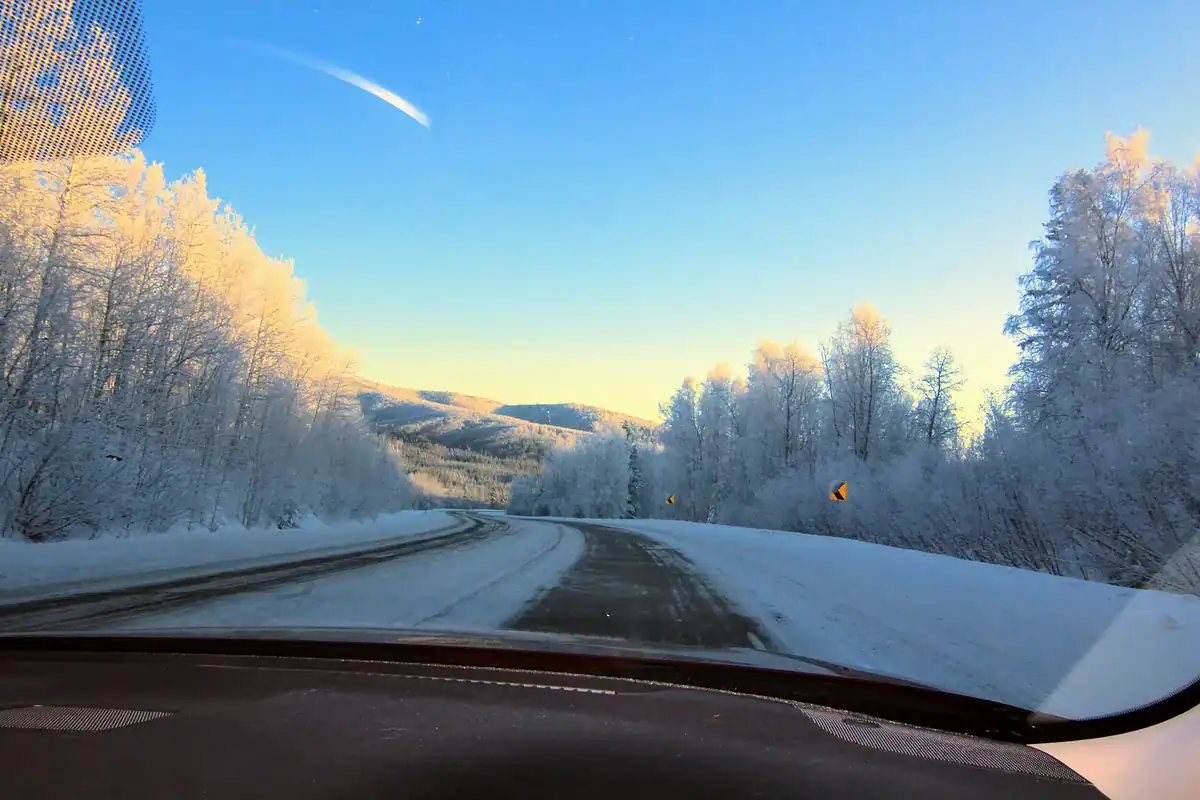 View from a car driving on a snowy, winding road lined with frosty trees under a clear blue sky and bright sunlight. Distant hills are visible, illuminated by the sun.
