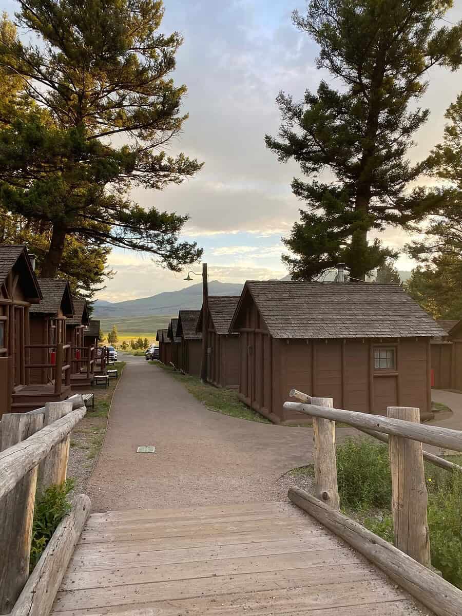 Wooden bridge leading to a row of rustic cabins surrounded by tall pine trees, with mountains visible in the distance under a partly cloudy sky at sunset.