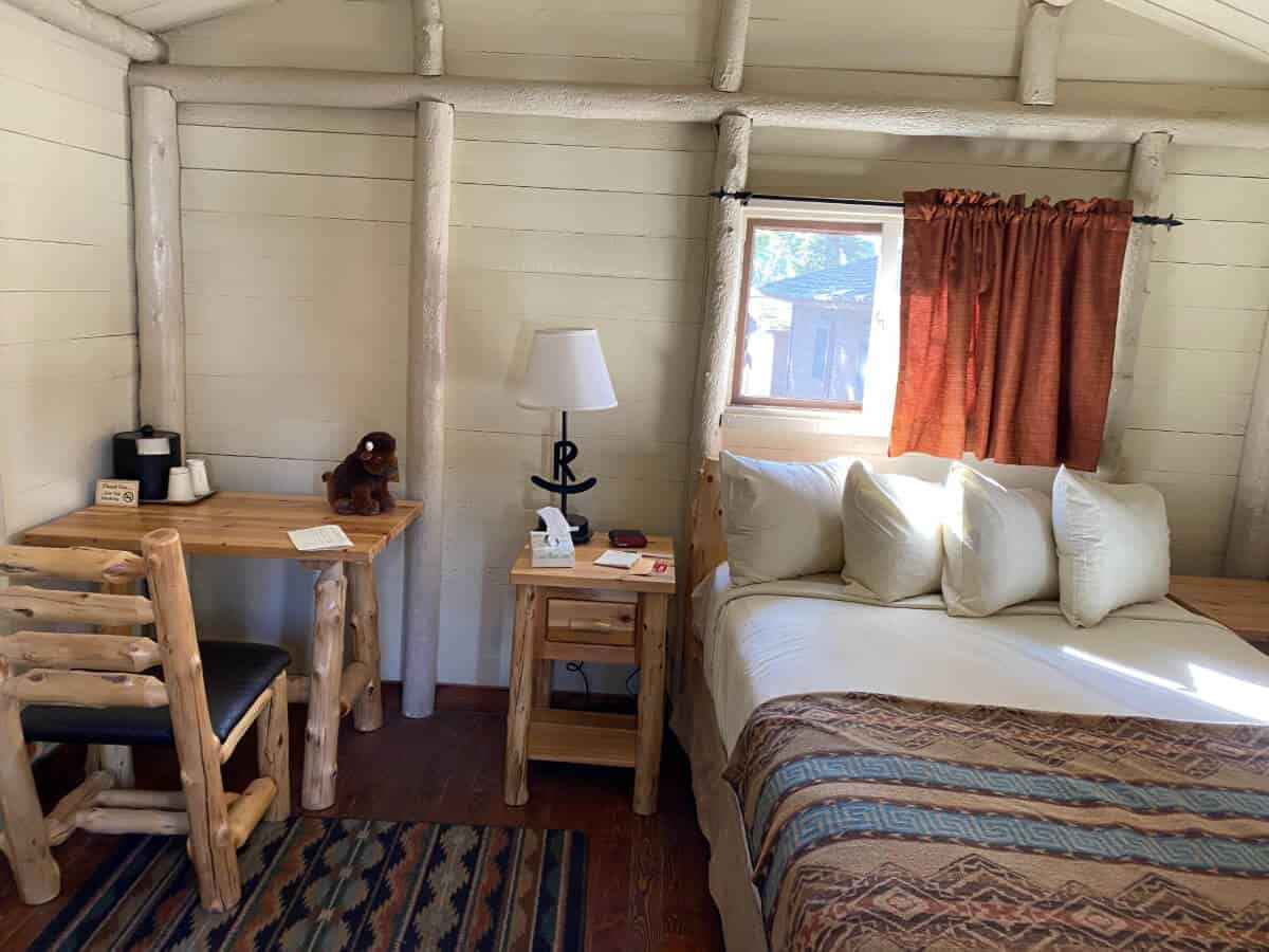 A cozy cabin room featuring a bed with a patterned quilt and white pillows, a wooden nightstand with a lamp, and a desk with a chair and a stuffed animal.