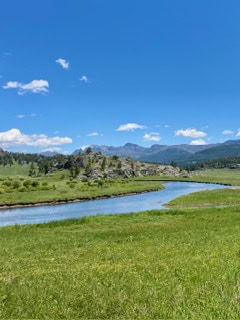 A winding river flows through a green meadow under a bright blue sky with scattered clouds and distant mountains in the background.