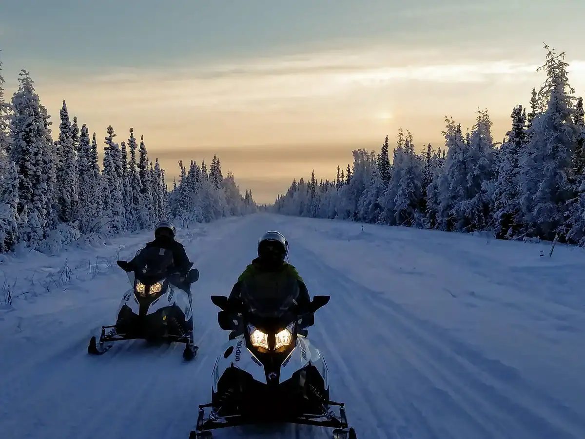 Two people ride snowmobiles down a snowy forest road at sunset. Snow-covered trees line both sides of the trail beneath a partly cloudy sky with soft, golden light.