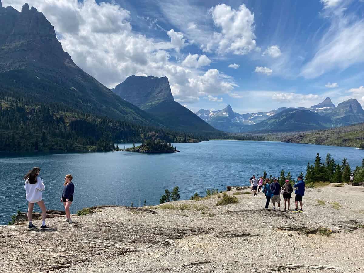 People stand and walk on a rocky overlook near a lake surrounded by forested mountains under a partly cloudy sky. Several tall peaks and distant snow-capped mountains are visible in the background.