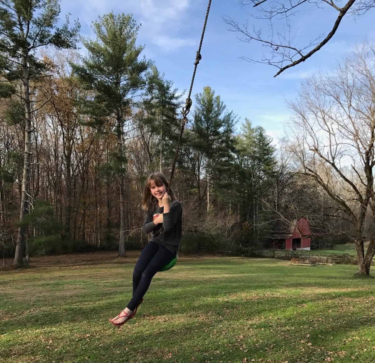 A young girl joyfully swings on a rope swing, an outdoor gift idea for kids, in a grassy field surrounded by tall trees. A red barn is visible in the background under the clear blue sky.