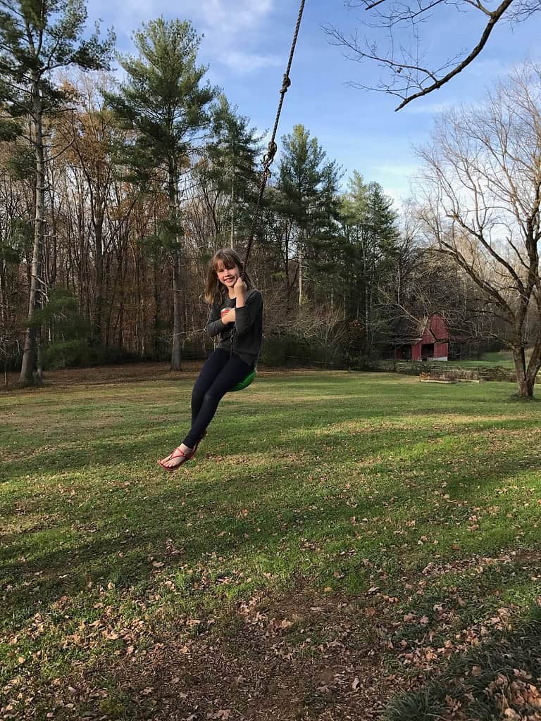 A person swinging on a rope swing in a grassy yard surrounded by trees, reminiscent of a serene stop on a Sedona itinerary. A red barn is visible in the background under a clear blue sky.
