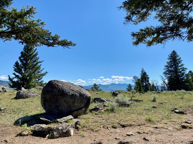 A large boulder sits on a grassy hill surrounded by trees, with distant mountains and a blue sky with scattered clouds in the background.