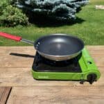 A frying pan with a red handle sits on a green portable Eureka! camping stove on a wooden table outdoors, with green grass, bushes, and a tree in the background under sunny skies.