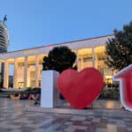 A large "I ❤️ T" sculpture stands in front of the illuminated Opera building in Tirana, Albania, at dusk, with modern and traditional architecture visible in the background.