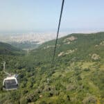 A cable car travels above a lush green mountain landscape, heading towards a distant city visible on the horizon under a clear blue sky.