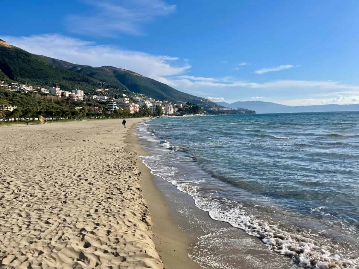 A sandy beach with gentle waves meets a cityscape at the base of green hills under a blue sky. A few people walk along the shore. Buildings and mountains are visible in the background.