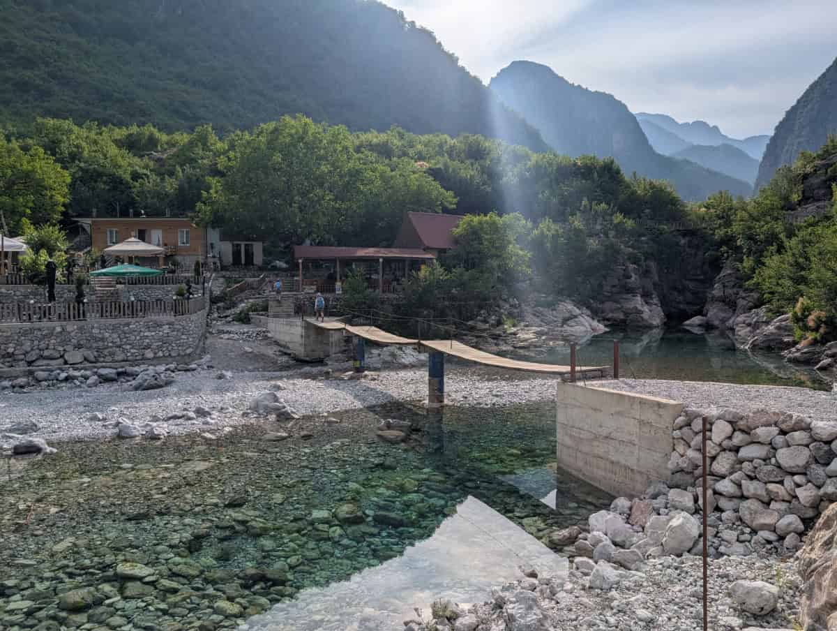 A narrow wooden footbridge spans a clear, rocky river with mountain peaks and lush green trees in the background. Nearby are small buildings and people enjoying the riverside scenery. Sunlight streams through the mountains.
