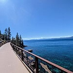 A paved pathway lined with railings stretches along a rocky shoreline, bordered by pine trees on the left and clear blue lake water on the right, beneath a bright, clear sky.