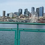 Seattle skyline with modern skyscrapers, the Seattle Great Wheel Ferris wheel, and waterfront buildings, viewed from behind a green railing over the water under a cloudy sky.