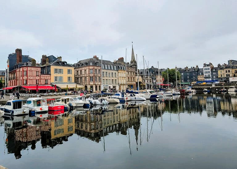 Colorful historic buildings and cafes line a waterfront marina with several docked boats reflecting in the calm water under a cloudy sky.