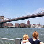 Two children stand by a railing, looking across the river at the Brooklyn Bridge and Manhattan skyline on a sunny day.