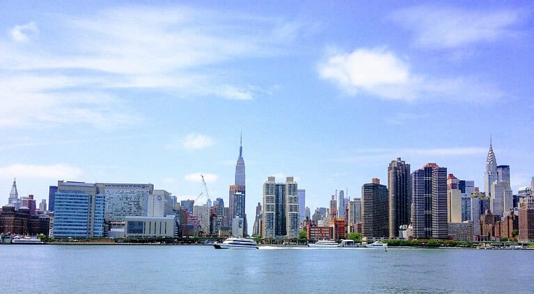 A view of the New York City skyline from across the river, showing tall buildings, boats in the water, and a blue sky with scattered clouds.