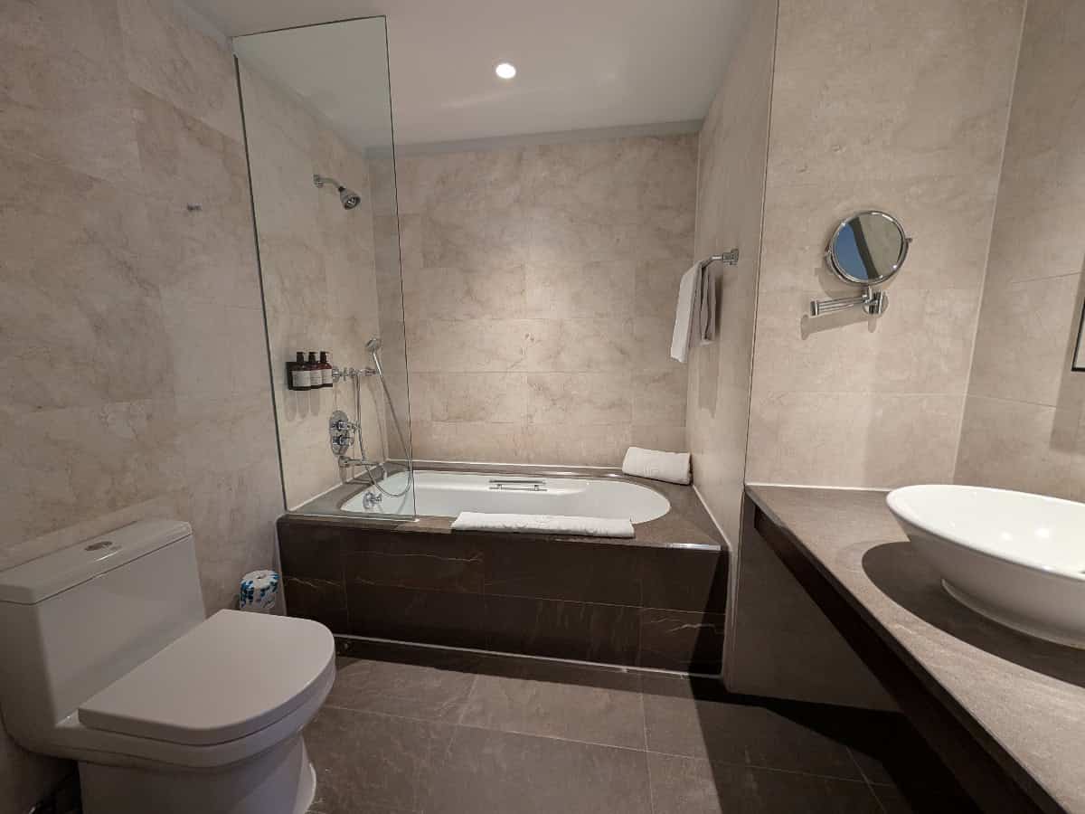 Modern bathroom with a toilet, bathtub with glass partition, wall-mounted shower, towels, sink with vessel basin, large mirror, and beige tiled walls. Bottles are placed on the bathtub ledge.