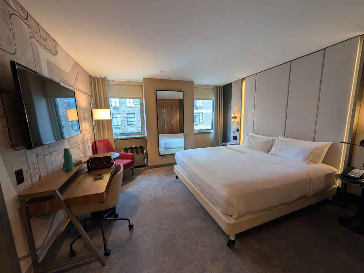 Modern hotel room with a large bed, desk, chair, TV on the wall, a red armchair by the window, a tall mirror, and natural light from two windows. The decor is neutral and contemporary.