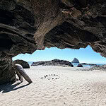 Oregon Coast Itinerary stop at Tunnel Beach. View from inside a rocky cave looking out onto a sandy beach, with rock formations and a large sea stack visible in the background under a clear blue sky.